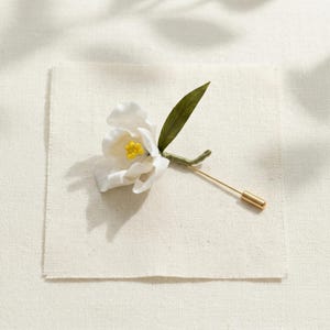 May include: A white flower lapel pin with a yellow center and a green stem and leaf. The pin has a gold-colored straight pin. The flower is set on a square of off-white fabric.