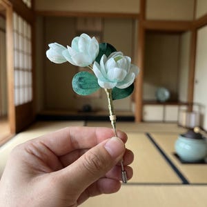 May include: A delicate, handmade floral lapel pin featuring three pale blue blossoms with green leaves. The pin has a gold-colored stem and a silver-toned pin backing. The flowers are held in a hand against a blurred background.