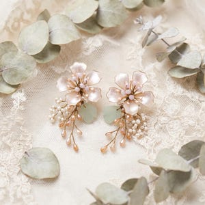May include: A pair of floral earrings featuring pale pink petals, small white seed beads, and light peach teardrop crystals arranged on gold-toned wire. Each earring includes a single soft green leaf accent, creating a delicate botanical design.