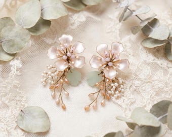 May include: A pair of floral earrings featuring pale pink petals, small white seed beads, and light peach teardrop crystals arranged on gold-toned wire. Each earring includes a single soft green leaf accent, creating a delicate botanical design.