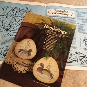 May include: A book titled "Renderings" featuring a collection of creative expressions in decorative painting techniques. The book cover features a painting of a pair of ducks in a pond setting. The book is open to a page with a detailed illustration of a floral design.