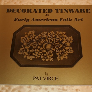 Puede incluir: Un libro titulado "Decorated Tinware: An Early American Folk Art" de Pat Virch. La portada es de color marrón dorado con texto negro y un diseño floral en el centro. El libro está abierto, mostrando la portada.
