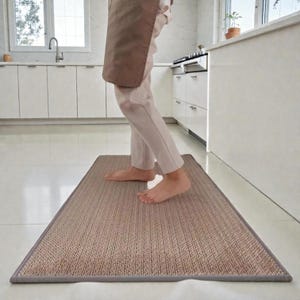Könnte beinhalten: Ein rechteckiger Küchenteppich mit gewebter Textur in Beige- und Brauntönen. Der Teppich hat einen grauen Rand und liegt auf einem weißen Fliesenboden. Das Bild zeigt eine Person, die in einer Küche auf dem Teppich steht.
