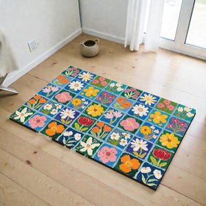May include: A rectangular floral rug with a grid pattern of colorful flowers on a blue background. The flowers are in shades of pink, yellow, orange, and white. The rug is placed on a wooden floor.