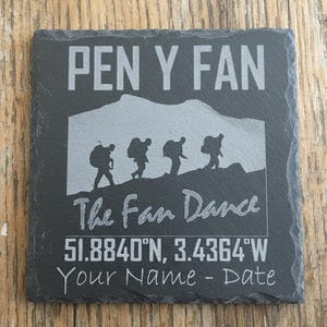 以下が含まれることがあります： 上部に「PEN Y FAN」の文字が入った、正方形の濃い灰色のスレートコースター。下にはハイカーのイラスト、「The Fan Dance」の文字、座標、パーソナライズ用のスペースがあります。