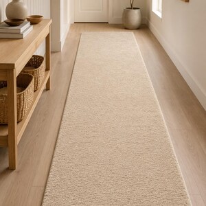 May include: A long, rectangular runner rug in a solid beige color with a textured, plush pile surface. The rug features clean, finished edges and is positioned lengthwise along the floor, extending from the foreground toward the back of the space.
