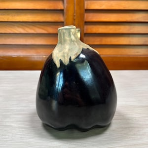 May include: A decorative ceramic eggplant figurine featuring a glossy, deep purple body and a textured, light beige stem. The object has a rounded, bulbous shape with a smooth, reflective surface finish and a distinct, realistic vegetable form.