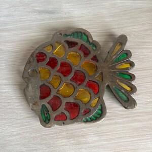 Puede incluir: Un objeto decorativo con forma de pez con un diseño de vidriera. El pez tiene un marco de metal plateado y está relleno de piezas de vidrio rojo, amarillo y verde. La cola y las aletas también son de metal plateado.