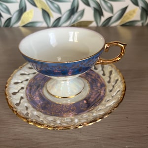 May include: A vintage teacup and saucer set. The teacup has a blue rim with gold accents and a gold handle. The saucer is white with a pierced design and a purple and gold center. A classic tea set.
