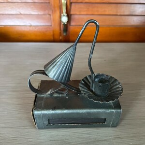 May include: A dark gray metal candle holder featuring a rectangular base, a small circular drip tray with a scalloped edge, and a curved handle attached to a conical snuffer. The metal has a matte finish with visible signs of wear and paint chipping.