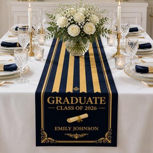 May include: A formal dining table set for a graduation celebration. The table is covered with a white tablecloth and features a navy blue and gold striped table runner with the text "GRADUATE CLASS OF 2026". The table is set with gold-rimmed plates, navy napkins, and crystal glassware.