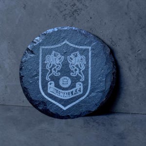 Peut inclure: Un dessous de verre rond en ardoise gris foncé avec un motif gravé au laser. Le motif représente un écusson avec deux créatures ressemblant à des lions et le texte "MILLWALL F.C." dans une bannière incurvée. Le dessous de verre a une surface rugueuse et texturée.