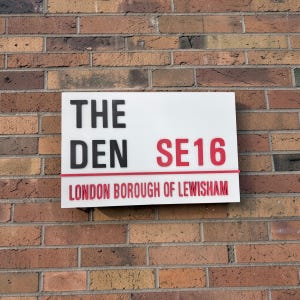 Peut inclure: Panneau rectangulaire blanc fixé sur un mur de briques. Le panneau affiche les mots "THE DEN" en noir, "SE16" en rouge et "LONDON BOROUGH OF LEWISHAM" en rouge. Il s'agit d'un panneau de signalisation.