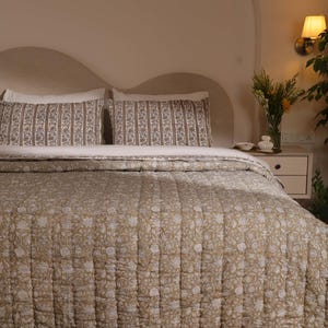 May include: A quilted bedspread and two matching rectangular pillow shams featuring a repeating floral pattern in muted tan and white. The bedding set is displayed on a bed with a curved headboard, showcasing a consistent block-print style design.