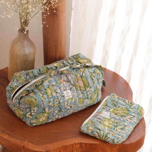 May include: A quilted rectangular toiletry bag and a matching flat zippered pouch, both featuring a light blue fabric with a green and white floral block print pattern. Each item includes a white zip and a small rectangular brand label.
