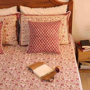 May include: A bed set featuring a quilt and matching pillow shams with a repeating floral pattern in shades of deep red and muted tan. A square throw pillow with a similar red floral print sits centred on the bed, with a notebook resting on top.