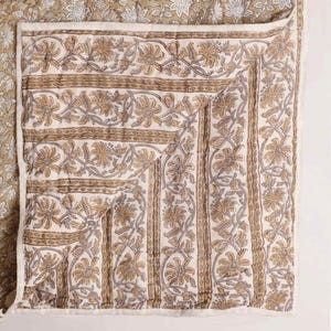 Puede incluir: Un textil acolchado con un estampado floral de bloques en tonos beige, marrón y blanco. La tela muestra un motivo repetitivo de flores y hojas estilizadas dispuestas en bandas verticales, con una esquina doblada que revela el reverso del tejido.