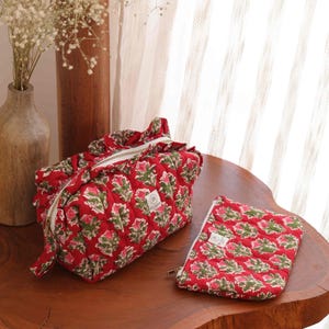 May include: A quilted fabric toiletry bag and a matching flat zippered pouch, both featuring a vibrant red background with a repeating green and pink floral block print pattern, white zipper closures, and a small rectangular brand label on each.