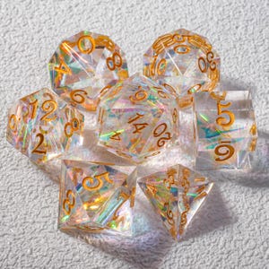 May include: A set of seven clear, iridescent polyhedral dice with gold-colored numbering. The dice are various shapes, including a d4, d6, d8, d10, d12, d20, and a percentile die. The dice are arranged on a textured white surface.
