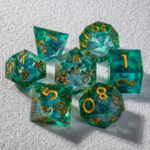 Op de afbeelding: Een set van zeven polyhedrale dobbelstenen in verschillende vormen. De dobbelstenen zijn transparant teal met goudkleurige cijfers en glitter. De dobbelstenen zijn ontworpen voor tabletop rollenspellen.