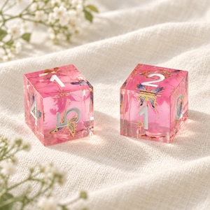 May include: Two translucent pink cubic dice featuring embedded butterfly graphics and gold-colored star accents. The dice have white engraved numbers on each face, including visible numerals 1, 2, 4, 5, and 8, with a smooth, polished finish.