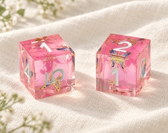 May include: Two translucent pink cubic dice featuring embedded butterfly graphics and gold-colored star accents. The dice have white engraved numbers on each face, including visible numerals 1, 2, 4, 5, and 8, with a smooth, polished finish.
