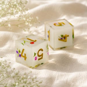 Puede incluir: Dos dados cúbicos de color blanco translúcido con pequeñas flores prensadas de colores y números dorados incrustados. Los dados muestran los números 1, 4, 5 y 7, con delicados detalles florales visibles en todo el material de resina.