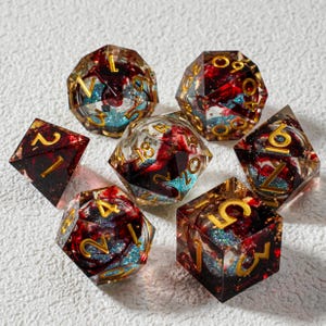 Puede incluir: Juego de siete dados poliédricos con un diseño único. Los dados son transparentes con detalles rojos, azules y dorados. Cada dado tiene números dorados. El juego incluye un D4, D6, D8, D10, D12, D20 y un dado de porcentaje.