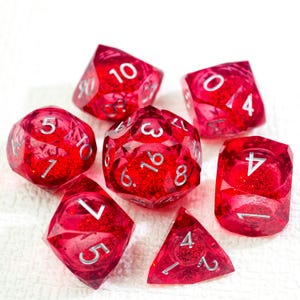 Puede incluir: Conjunto de siete dados poliédricos rojos con numeración plateada. Los dados son transparentes con purpurina en su interior. El conjunto incluye varias formas, como d4, d6, d8, d10, d12 y d20, comúnmente utilizados en juegos de rol.