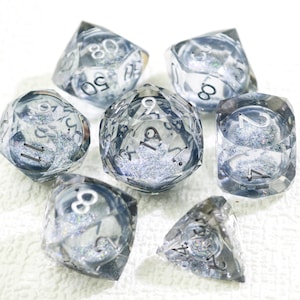 Può includere: Un set di sette dadi trasparenti e sfaccettati con glitter argentati all'interno. Ogni dado ha numeri bianchi. I dadi sono disposti in un motivo circolare, con varie forme e dimensioni, adatti per giochi da tavolo.