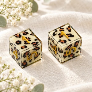 May include: Two square dice featuring a cream-colored base with a black and brown leopard print pattern. Each die face displays a raised, metallic gold number, including the numerals one, four, and five, set against the animal print design.