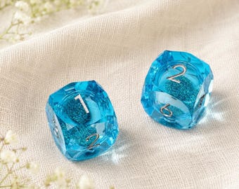 May include: Two translucent blue six-sided dice featuring silver-coloured numbers. Each die contains embedded blue glitter particles within the clear resin material. The numbers one, two, and six are clearly visible on the faceted surfaces of the dice.