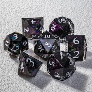 Può includere: Un set di sette dadi poliedrici di varie forme. I dadi sono neri con inclusioni iridescenti e numerazione bianca. Questi dadi sono progettati per giochi di ruolo da tavolo.