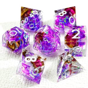 Peut inclure: Un ensemble de sept dés polyédriques avec un motif floral violet et rose. Les dés sont transparents avec des chiffres argentés. Les dés sont de formes variées, dont un d4, d6, d8, d10, d12 et d20.