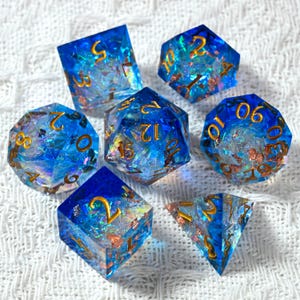 Puede incluir: Juego de siete dados poliédricos de varias formas. Los dados son de color azul translúcido con números dorados e incrustaciones de escamas iridiscentes y color cobre. Los dados están diseñados para juegos de rol de mesa.