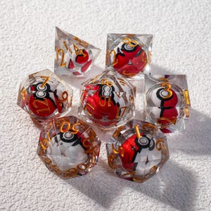 May include: Set of seven clear resin dice with gold numbering. Each die features a red, white, and black design. The dice are in various geometric shapes, including a cube, a pyramid, and a multi-sided die.