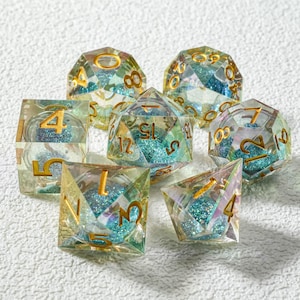 May include: A collection of clear, multi-sided dice with gold numbering and iridescent blue glitter filling. The dice are arranged on a white surface, showcasing their various shapes and sizes, perfect for tabletop games.