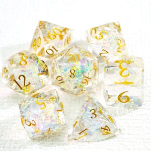 Può includere: Un set di sette dadi trasparenti e iridescenti con numeri dorati. I dadi hanno varie forme, tra cui un d20, d12, d10, d8, d6, d4 e un dado percentuale. I dadi sono progettati per giochi di ruolo da tavolo.