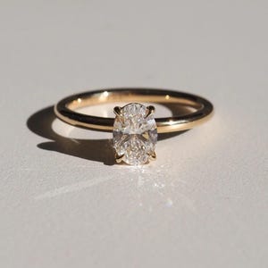 May include: A gold ring with a clear, oval-cut gemstone. The ring band is thin and polished, and the gemstone is set in a classic prong setting. The ring is displayed on a white surface.