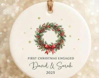 Könnte beinhalten: Runder, cremefarbener Weihnachtsschmuck mit einem Kranzdesign. Der Kranz zeigt Tannennadeln, rote Beeren, Tannenzapfen und eine rote Schleife. Der Text lautet "First Christmas Engaged" und "David & Sarah 2025". Ein brauner Faden ist befestigt.