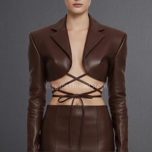 May include: A brown leather jacket with long sleeves and a cropped design. The jacket features a lapel collar and a crisscross tie closure at the waist. The bottom portion of the jacket is a matching brown leather skirt.