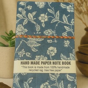 May include: A rectangular notebook with a blue cover featuring a white floral pattern. A multi-coloured string wraps around the centre. A white paper band across the bottom reads: HAND MADE PAPER NOTE BOOK "This book is made from 100% handmade recycled rag. tree free paper"