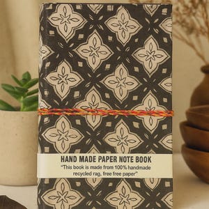 May include: A rectangular notebook with a black and beige geometric floral pattern. A white paper band across the center reads HAND MADE PAPER NOTE BOOK and This book is made from 100% handmade recycled rag, free free paper. A thin orange string wraps around.