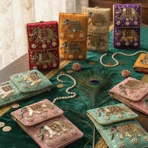 May include: An assortment of colorful, embroidered elephant-themed clutch purses and small wallets. The purses are in various colors, including red, orange, teal, pink, and purple, and are displayed on a green table runner.
