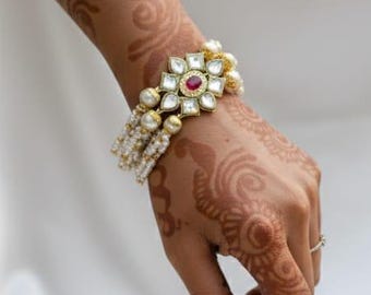 May include: A gold-toned bracelet with a central floral design, featuring a red gemstone and surrounded by white pearls. The bracelet is worn on a hand adorned with henna patterns, set against a white fabric backdrop.