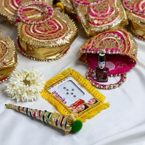 May include: An assortment of ornate, gold-colored gift boxes with pink and gold accents, along with matching bracelets. A small yellow frame with the name "anamika" is also present, along with a decorative cone and a white flower garland.