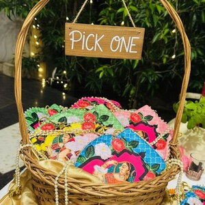 Può includere: Un cesto di vimini pieno di ventagli colorati e fantasia. I ventagli presentano disegni floreali e ritratti in rosa, blu, verde e giallo. Un cartello di legno con la scritta "PICK ONE" è appeso sopra il cesto.
