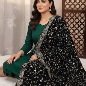 May include: A woman wearing a dark green dress with a black shawl. The shawl is decorated with an intricate gold and silver paisley pattern. The shawl is draped over the woman's shoulders and around her arms.