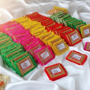 Puede incluir: Una colección de objetos decorativos cuadrados y coloridos. Cada objeto presenta un diseño central con puntos, enmarcado por un borde dorado y bordes con flecos en varios colores, incluyendo rojo, rosa, amarillo y verde. Los objetos están dispuestos sobre una superficie blanca y sedosa.