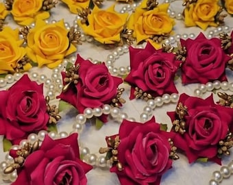 May include: An assortment of floral wrist corsages. The corsages feature vibrant red, yellow, and burgundy roses, accented with gold-colored embellishments and arranged on pearl bracelets. The background is a light-colored surface.
